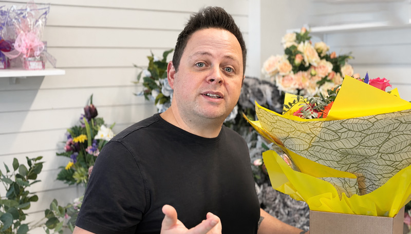 A man in a floristry shop holds a large yellow-wrapped bouquet in a box, speaking to camera,
