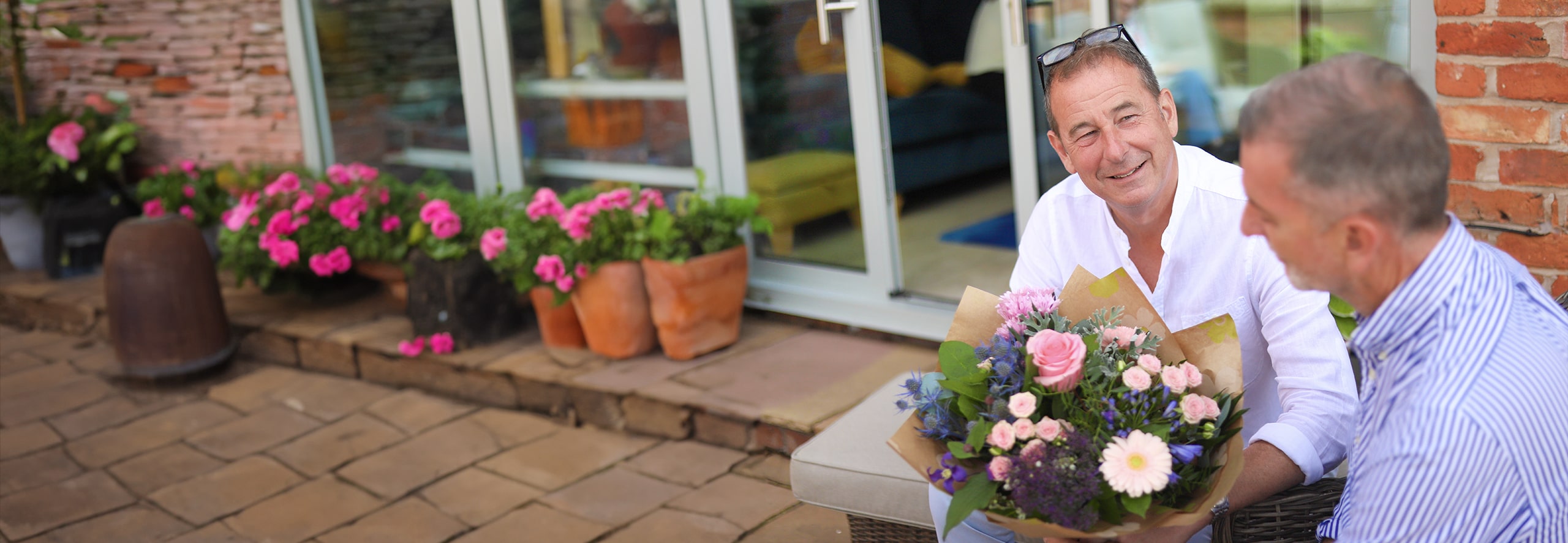 Man giving a freshly wrapped bouquet to a smiling friend on a garden patio.
