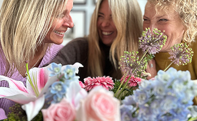 Three women smiling and laughing together behind a colourful bouquet of pink roses, blue hydrangeas, and lilies.