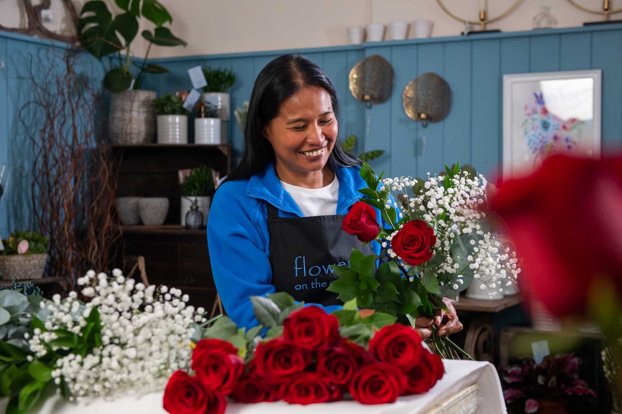 Happy florist inside shop making a red rose bouquet with flowers layed on worktop
