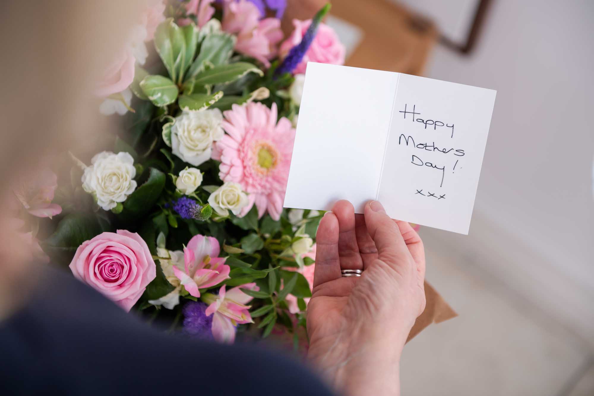 Bouquet in the background with a hand holding a gift card which says "Happy Mother's Day!"