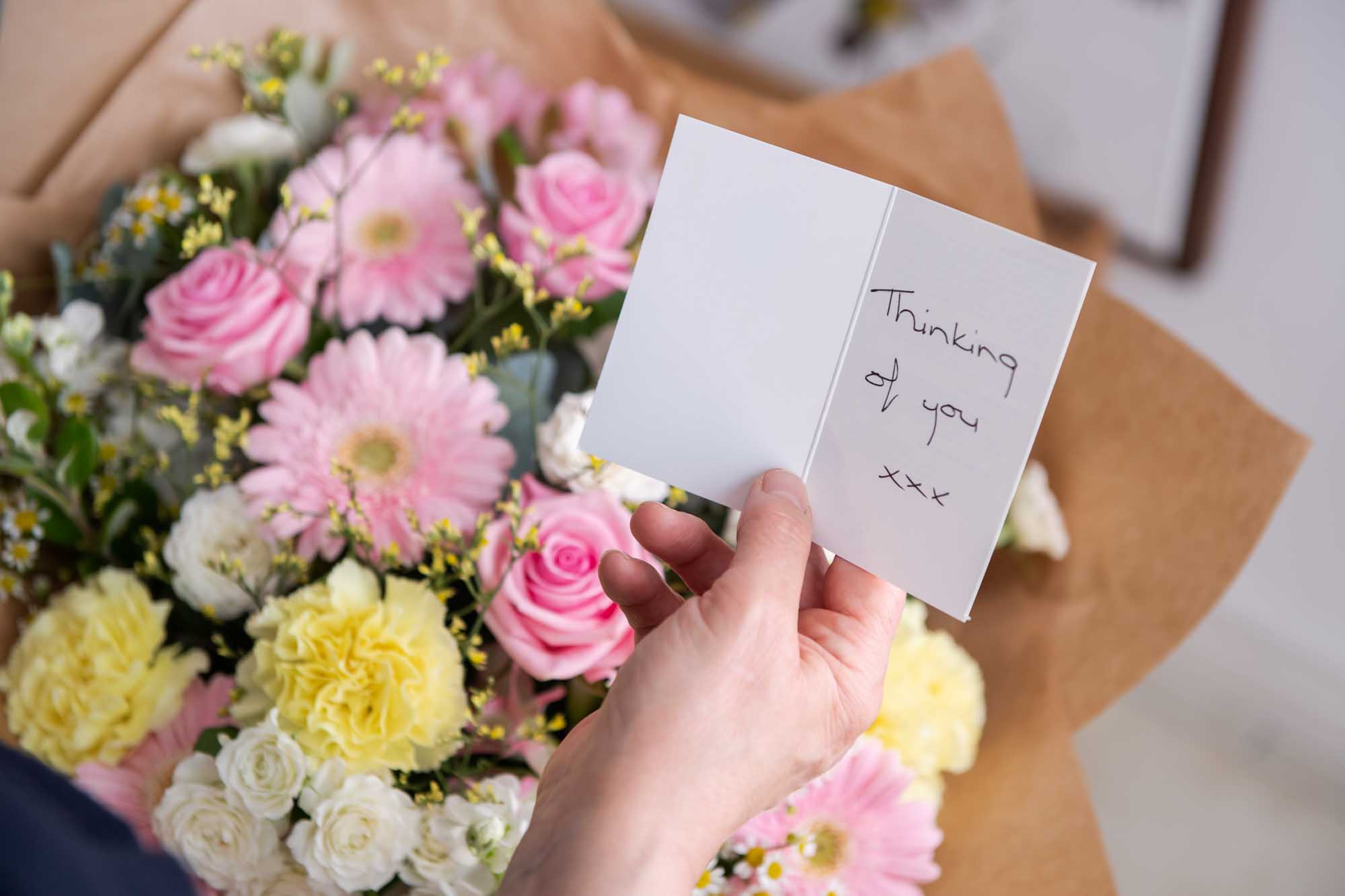 Hand holding a flower card the says "Thinking of you" with pink and yellow themed flowers in the background