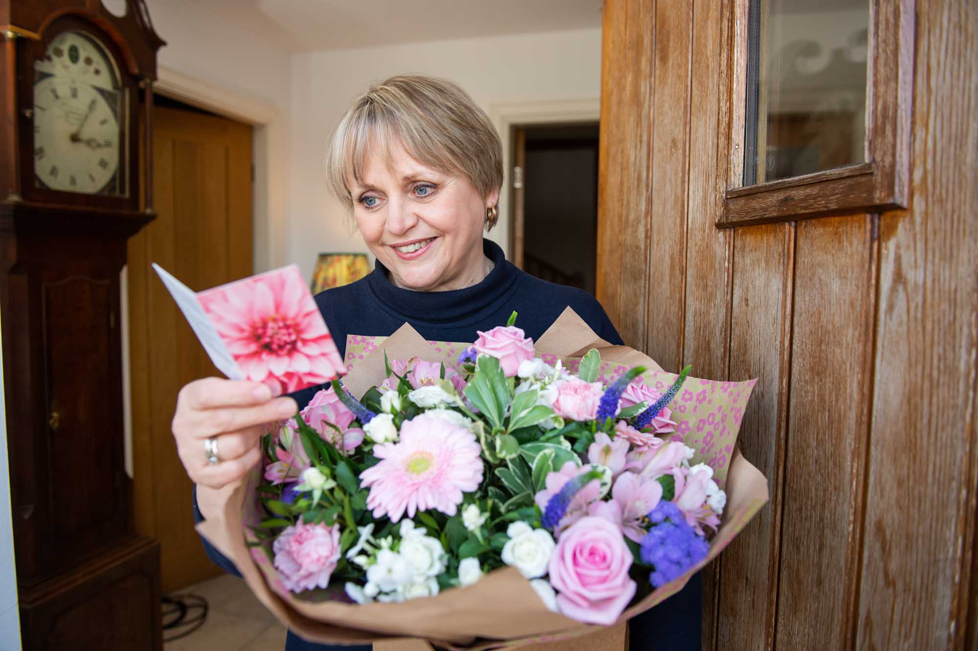 Smiling woman at front door reading a flower card whilst holding a pastel themed bouquet