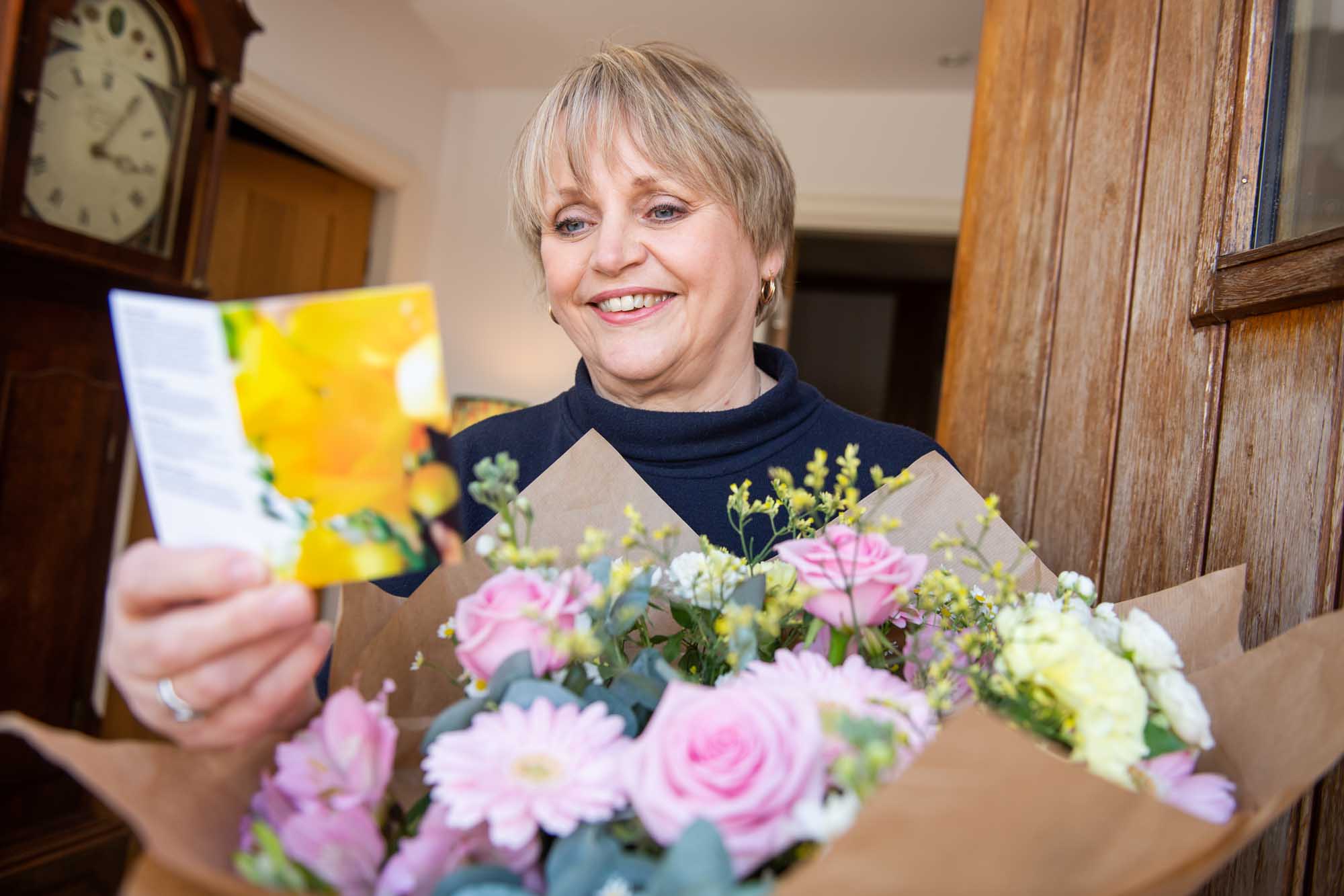 Smiling woman at front door reading a flower card with a bokeh of pastel flowers in the foreground