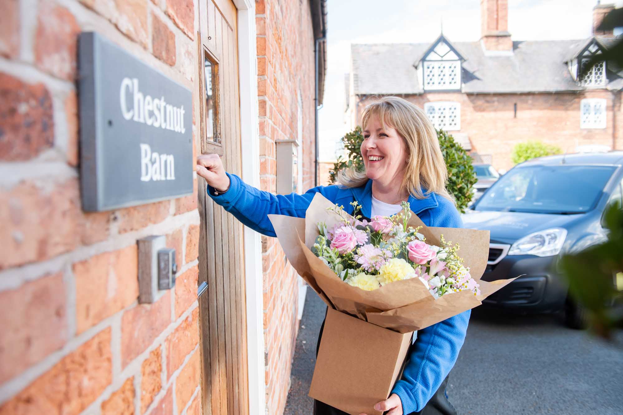 Smiling florist at front door holding a pastel themed Mother's Day Bouquet ringing the door bell