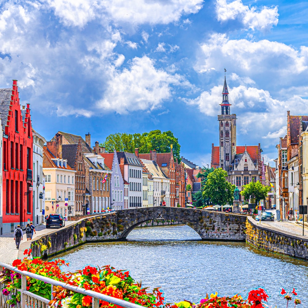 Bruges canal scene with colourful historic buildings, a stone bridge, and flowers.