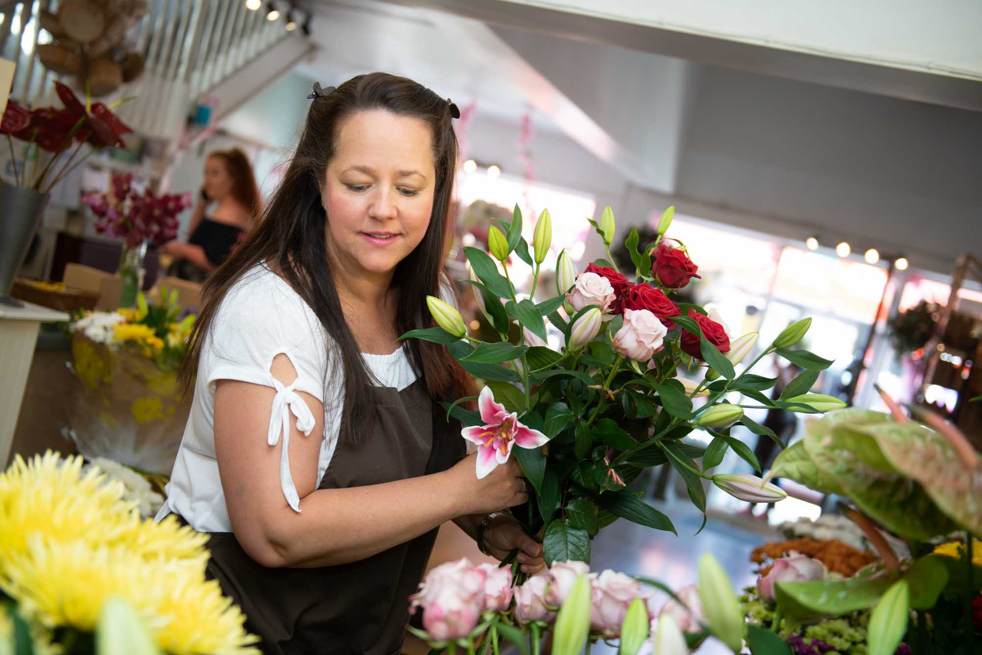 Florist in a brown apron holding a bouquet of red and pink roses with lilies inside a bright and welcoming flower shop.