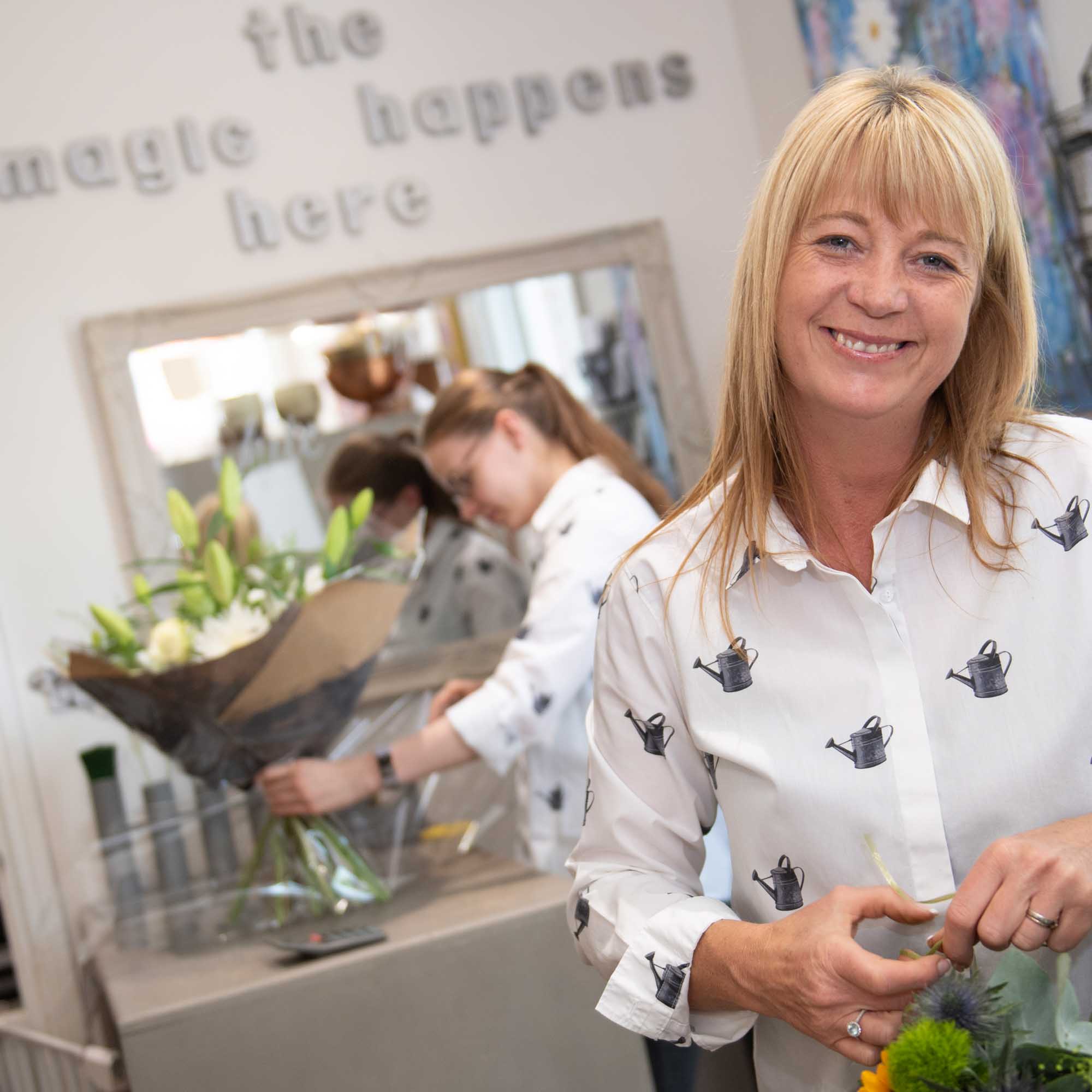 Florist wearing white shirt smiling with another florist in the same shirt making a lily bouquet in the background