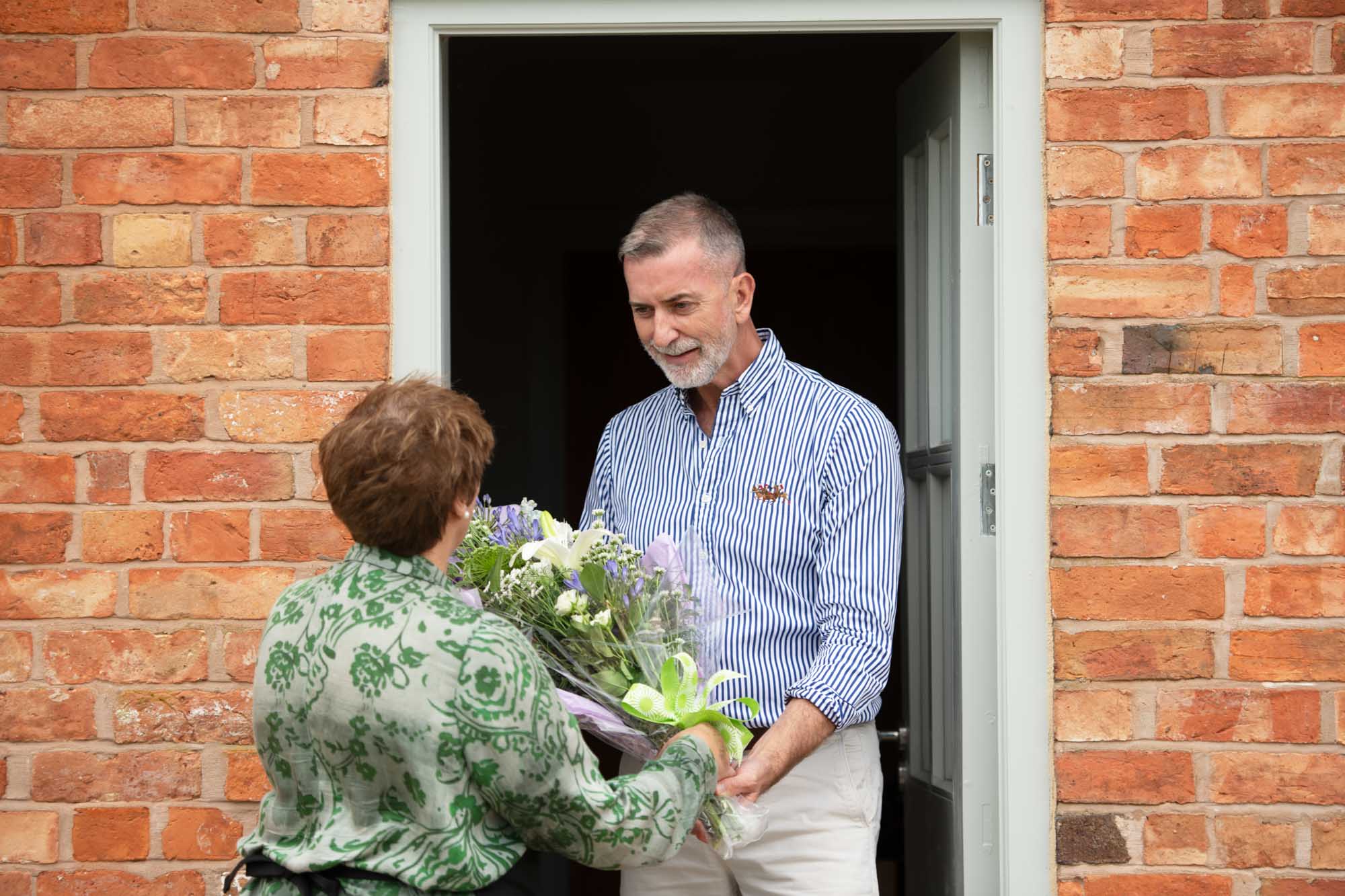 Man in a blue striped shirt at front door receiving a wrap of lilac and cream flowers from a florist