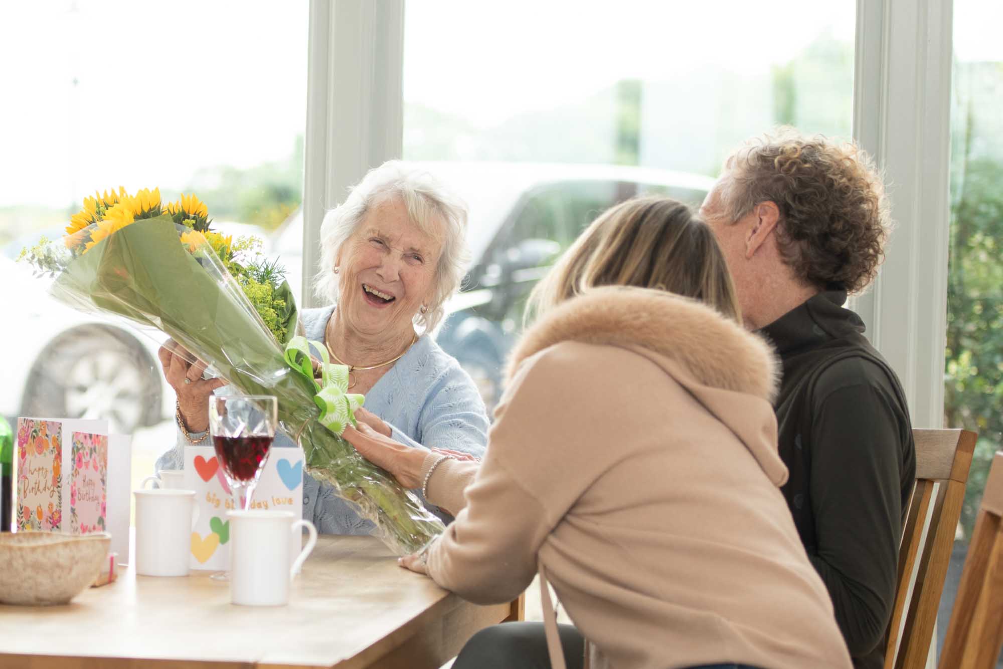 Elderly woman smiling as she receives a bouquet of sunflowers at a table with family.