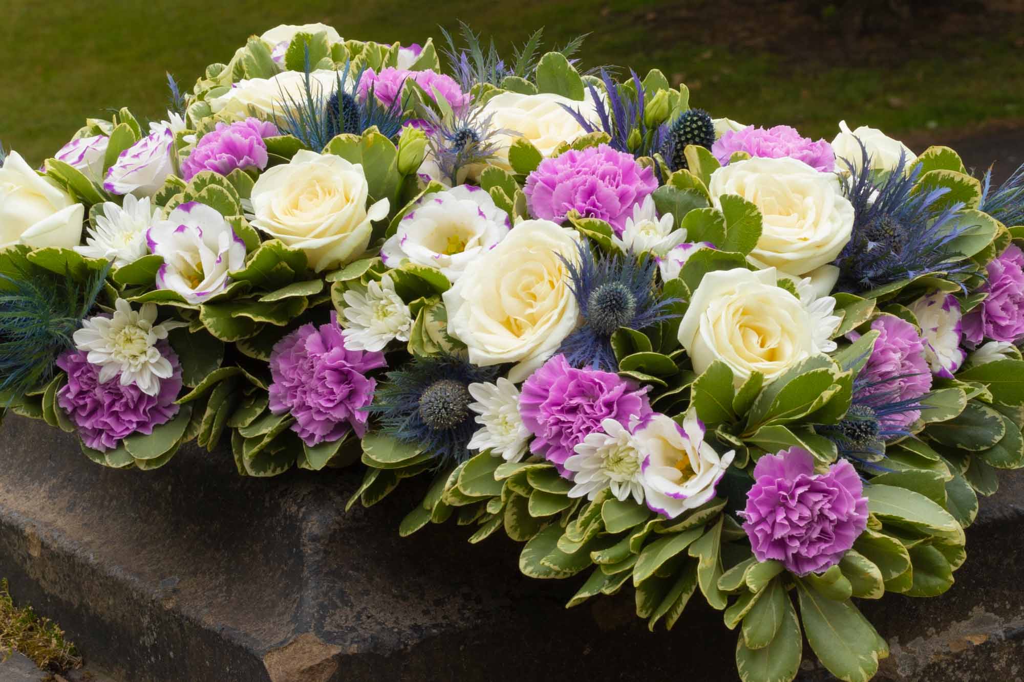 Crop of a funeral pillow in cream and purple flowers on stone ground with a green grass background.