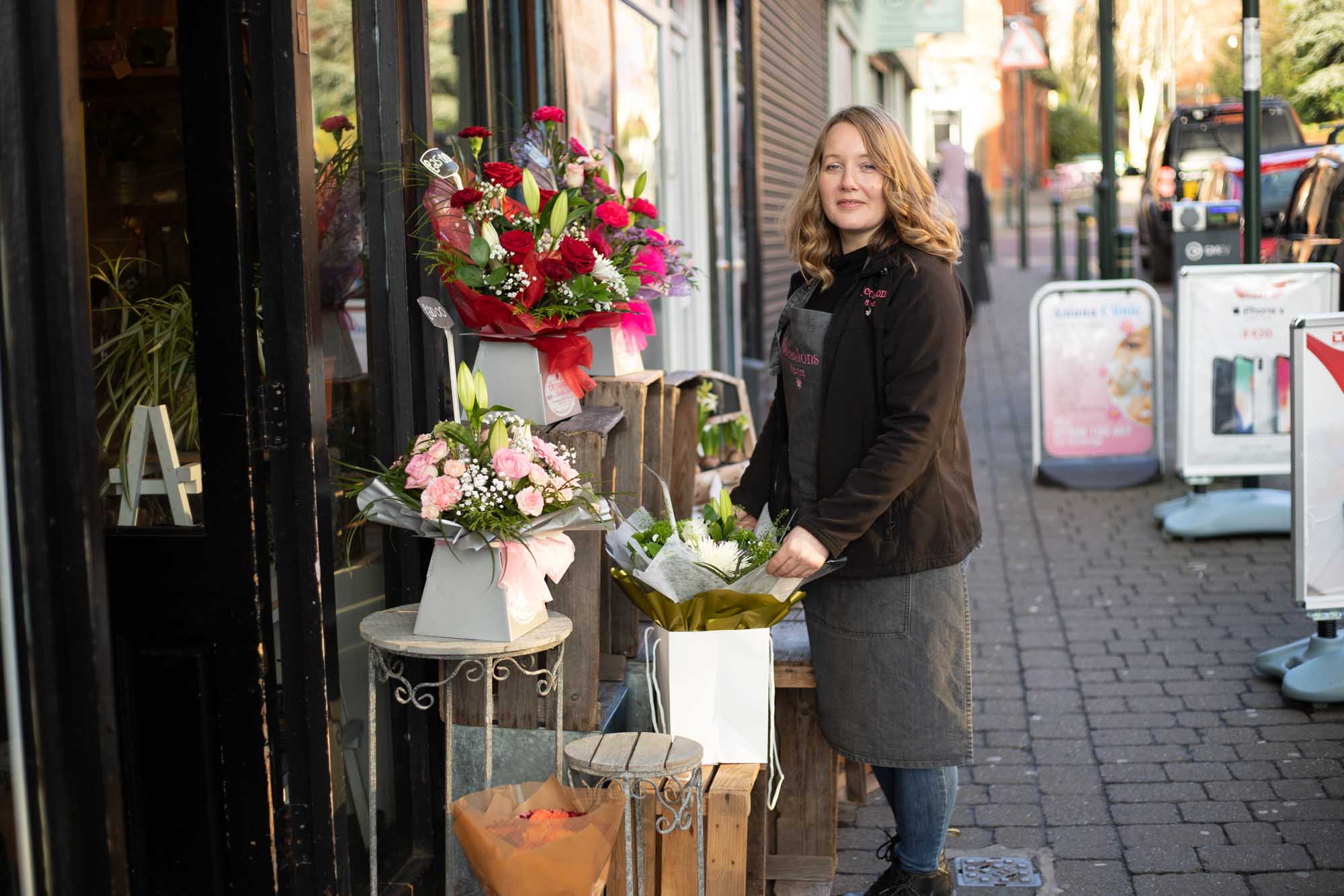 Florist standing outside a flower shop, holding a fresh bouquet, surrounded by beautifully arranged flower displays.