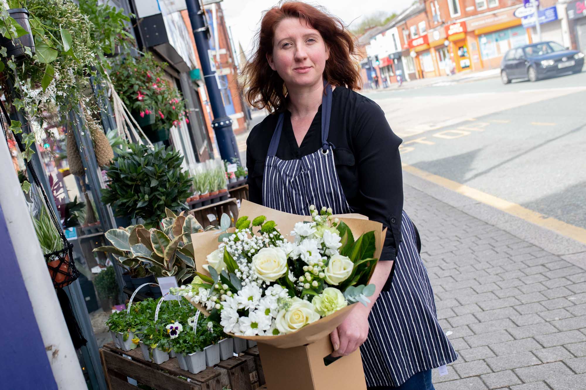 Florist in a striped apron holding a bouquet of white roses and fresh greenery outside a charming floral boutique.