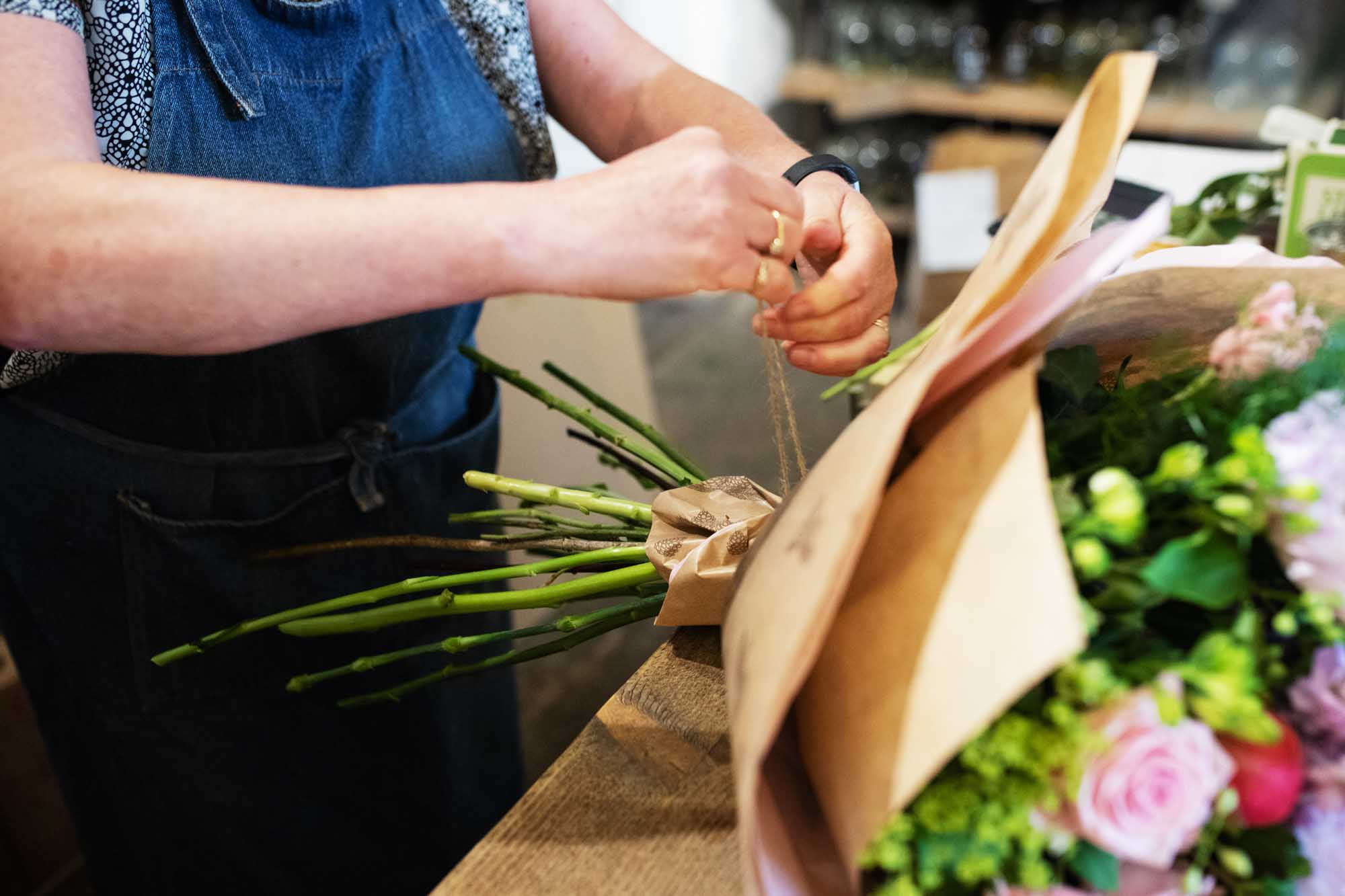 Florist carefully tying a ribbon around the stems of a freshly arranged bouquet on a wooden worktable.