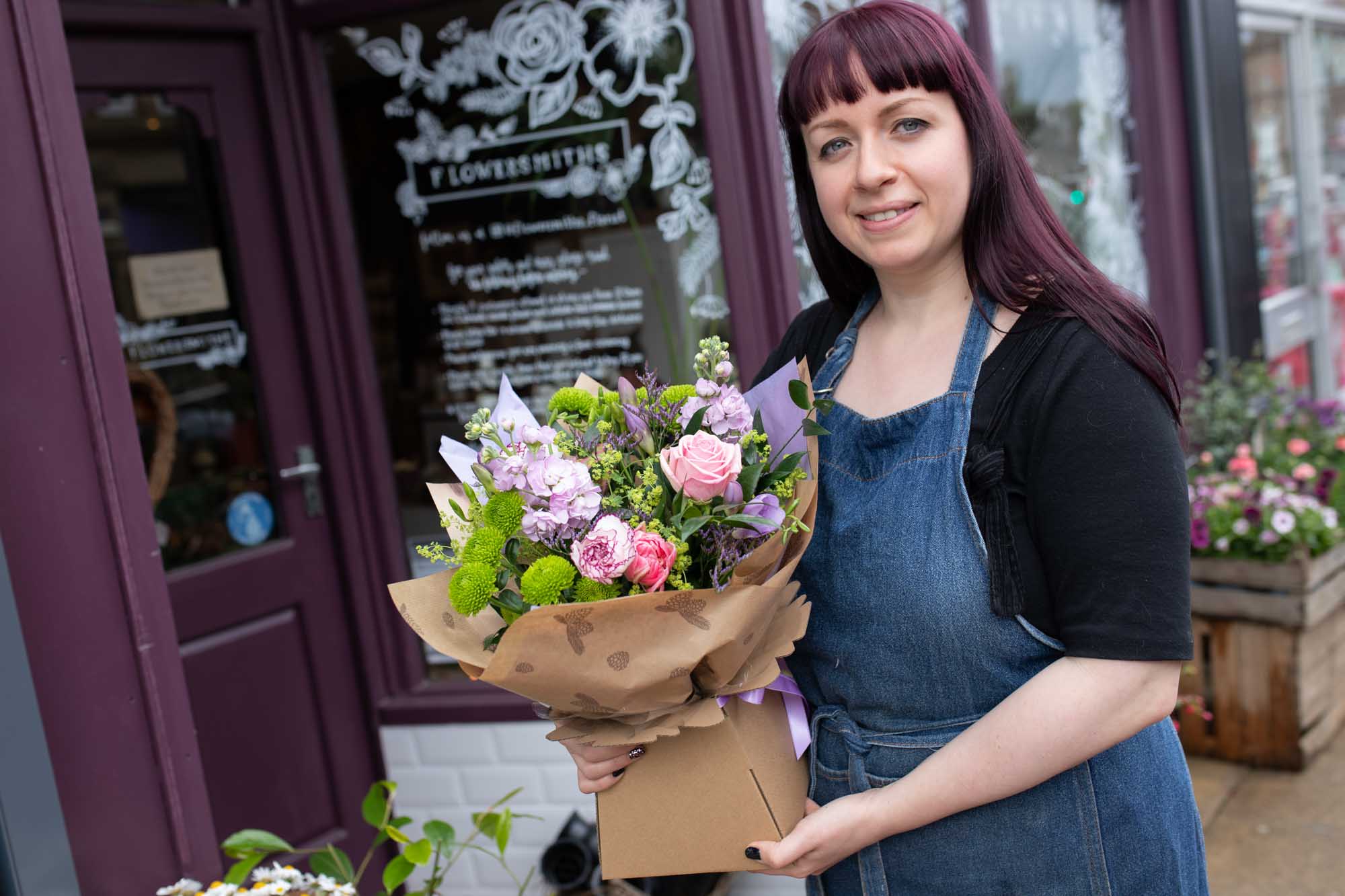 Smiling florist in a denim apron presenting a hand-tied bouquet of pink roses and lush greenery outside her shop.