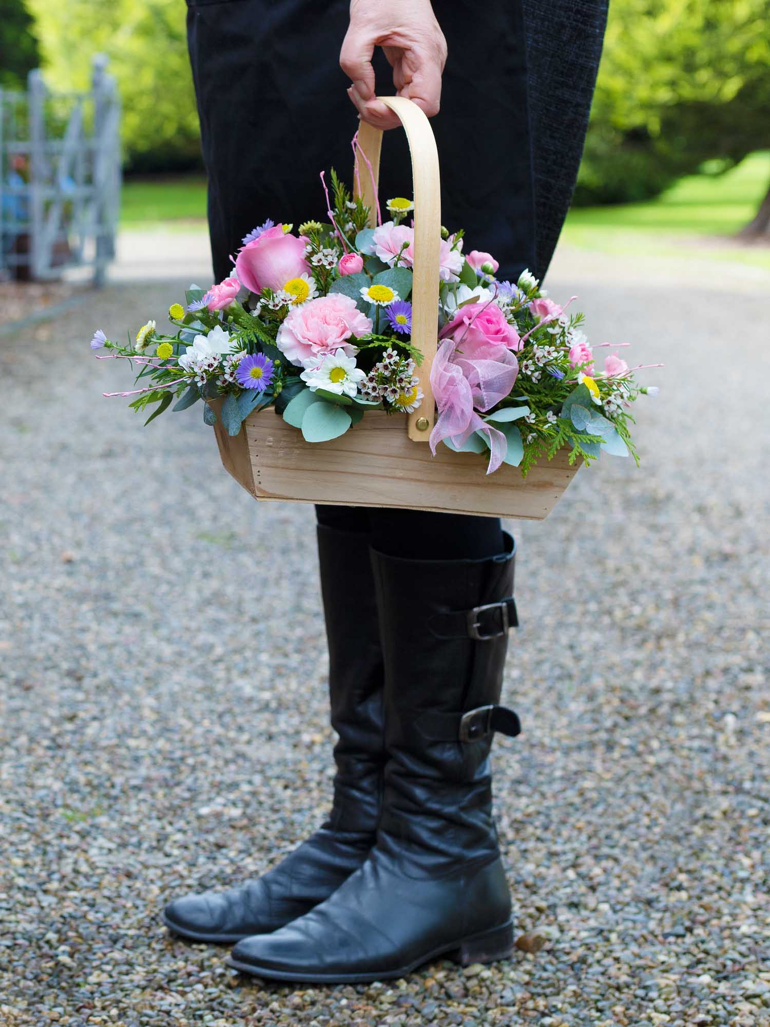 Person holding a wooden basket of flowers while wearing black boots on a gravel path