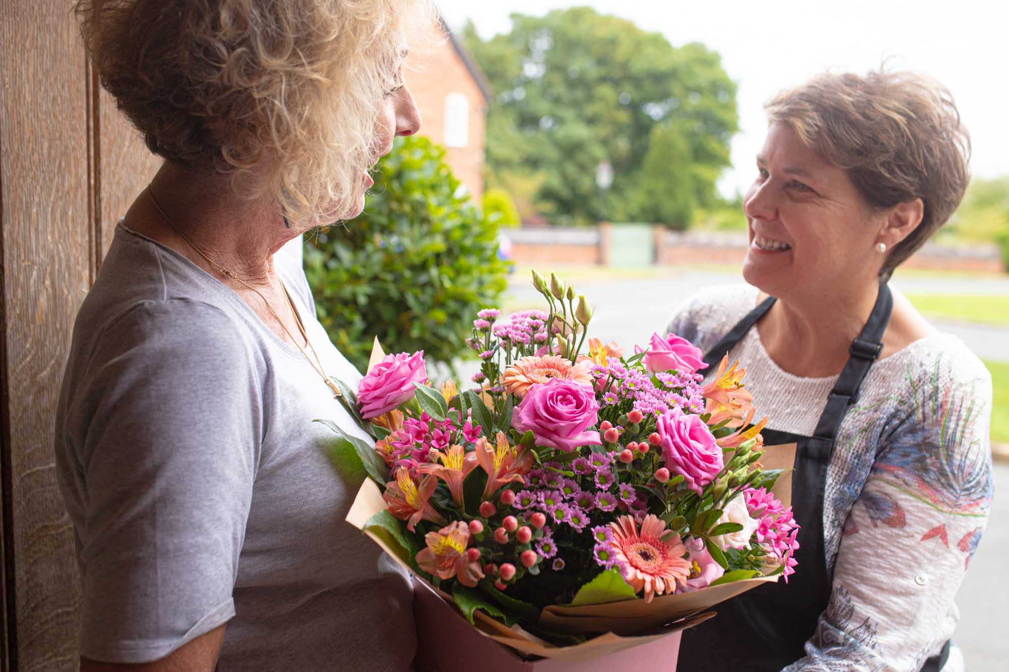 Smiling florist delivering a vibrant bouquet of pink and orange flowers to a happy recipient at their doorstep.