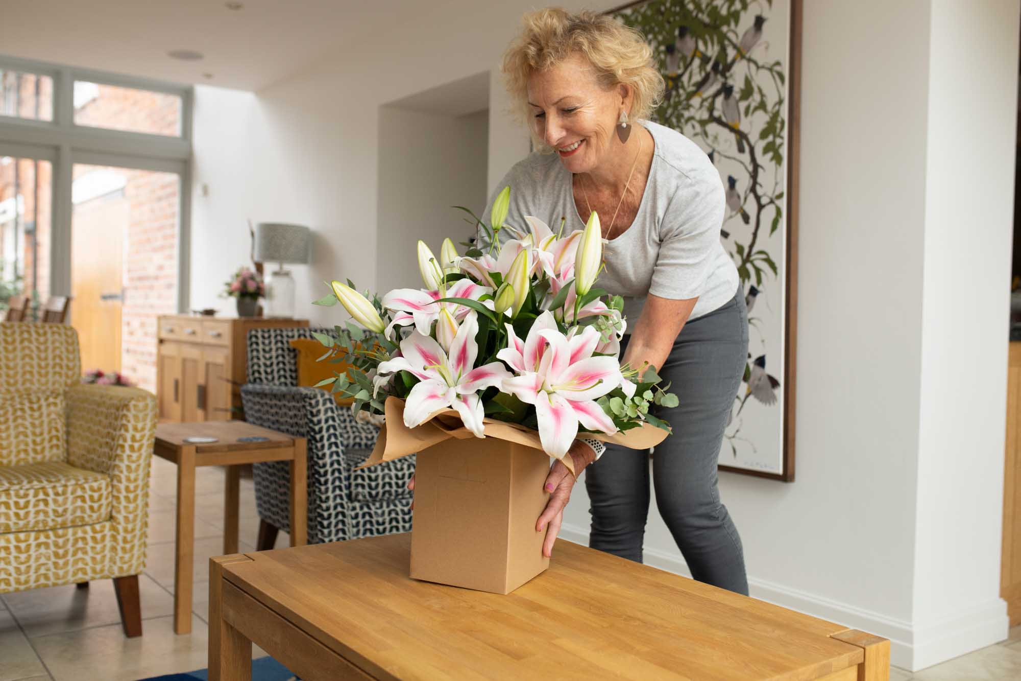 Woman in her home putting a box of oriental lilies on her coffee table smiling