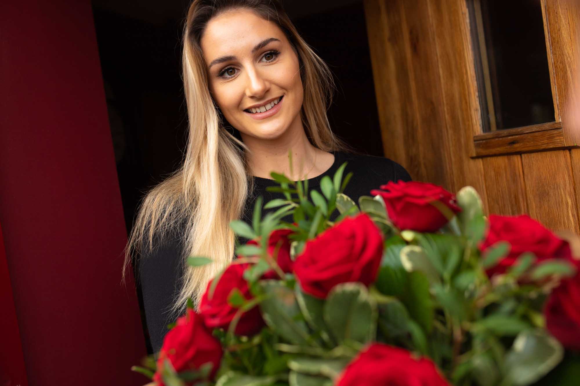 Woman smiling while holding a bouquet of red roses at the door.