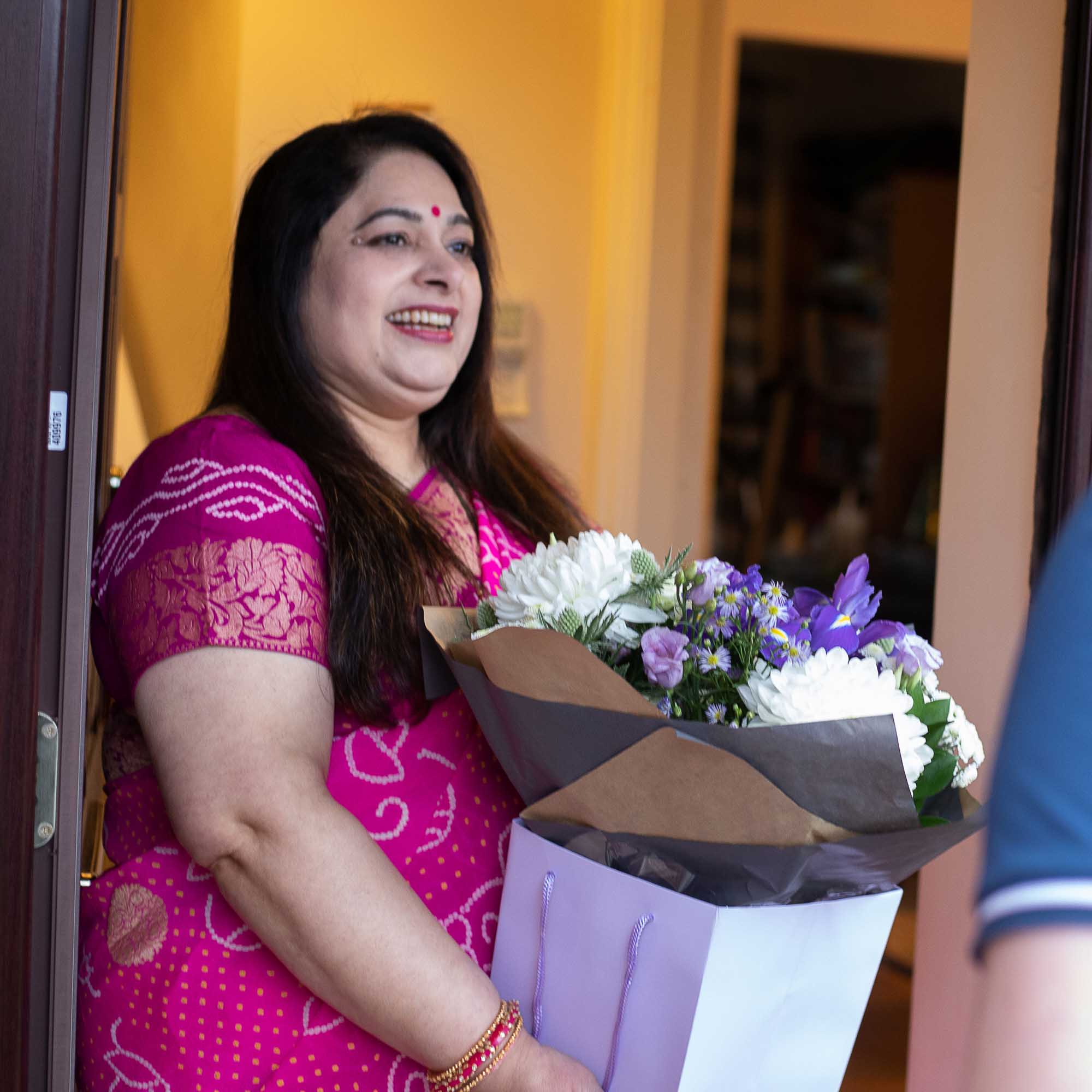 Happy indian women wearing a pink saree holding a purple themed box of flowers at her front door.