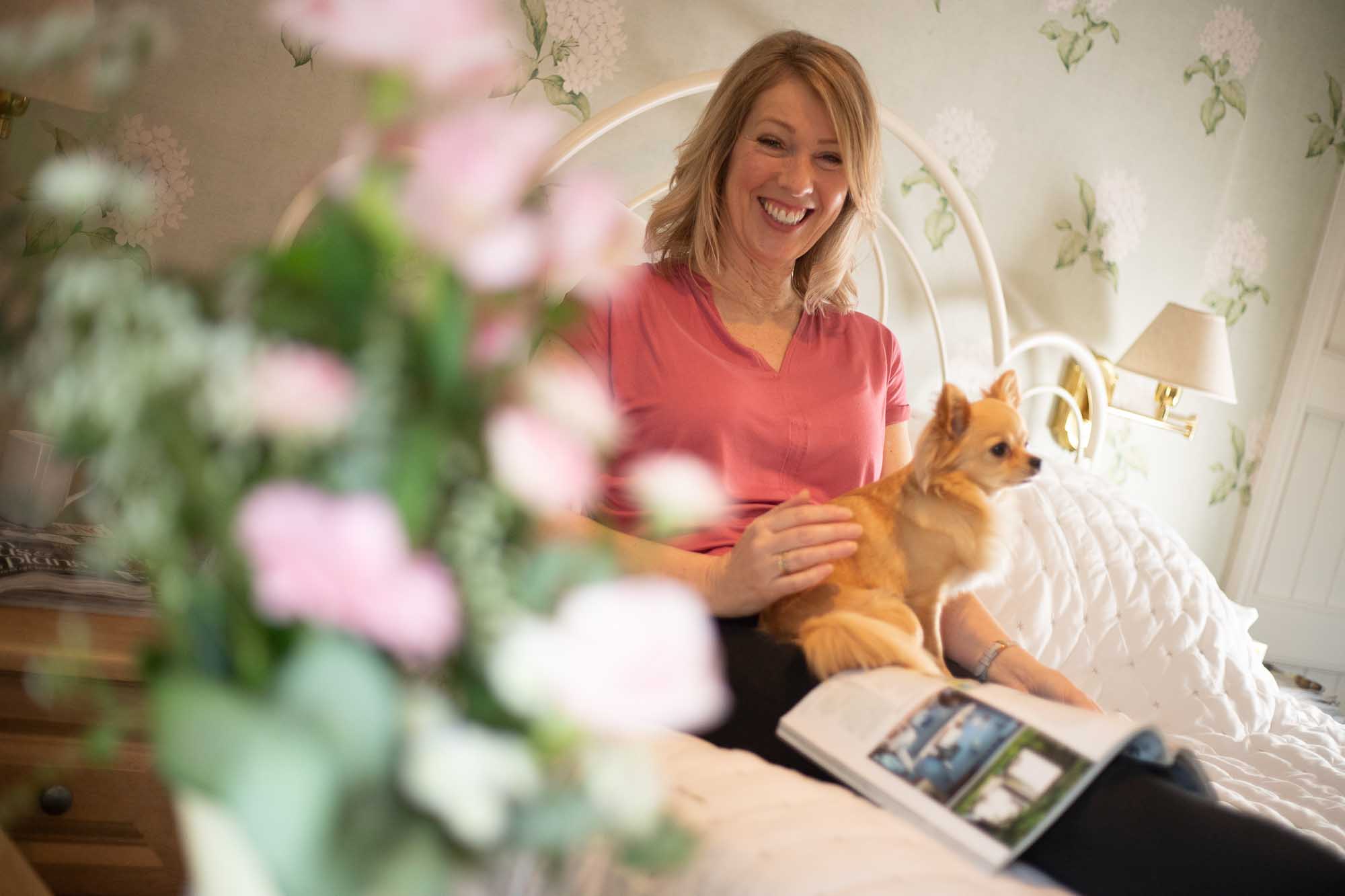 Happy woman and small dog in sat on bed with a magazine with a bokeh of pastel pink flowers in the foreground