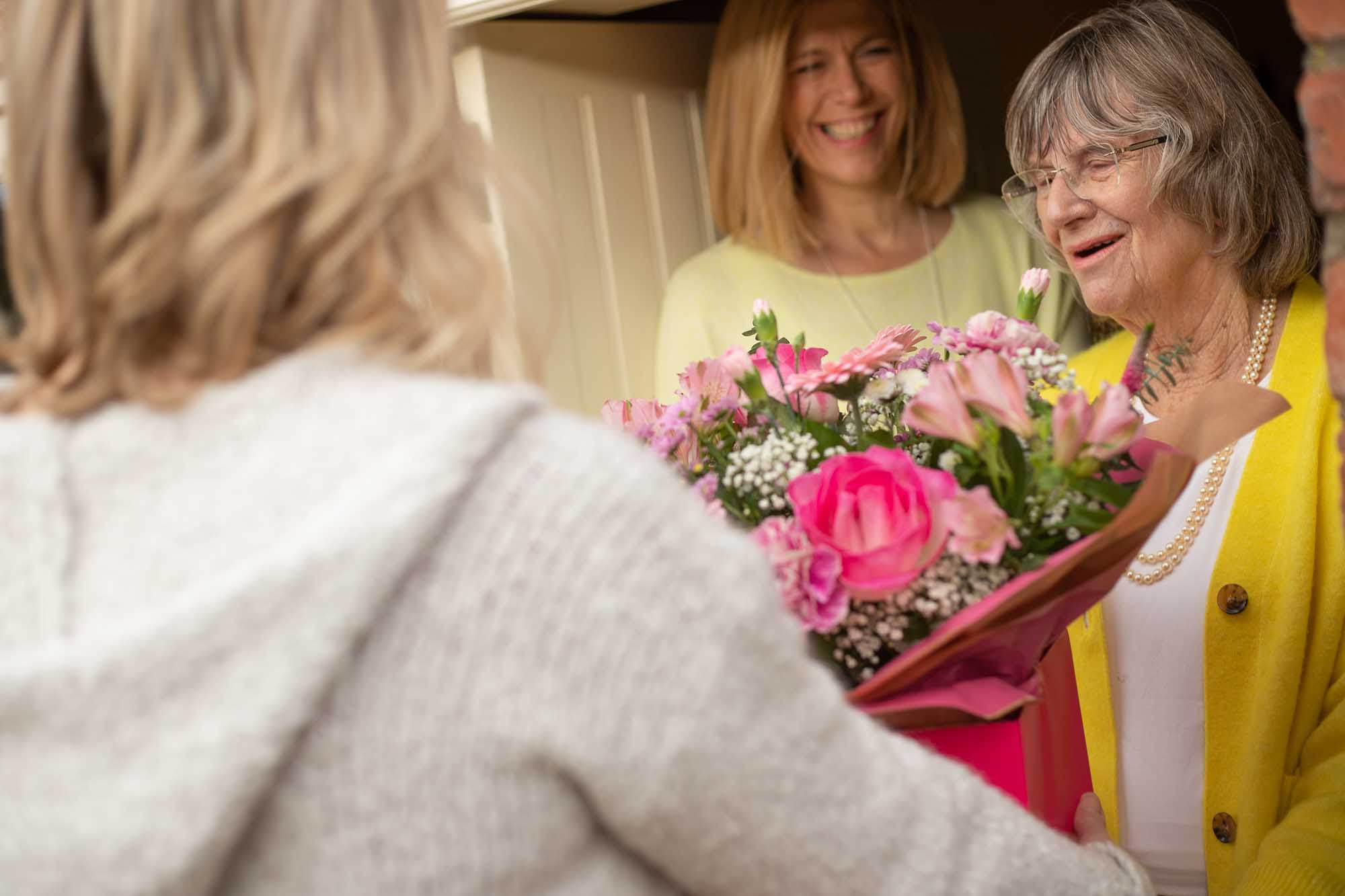 Elderly woman receiving a bouquet of pink flowers with a smile at her front door.