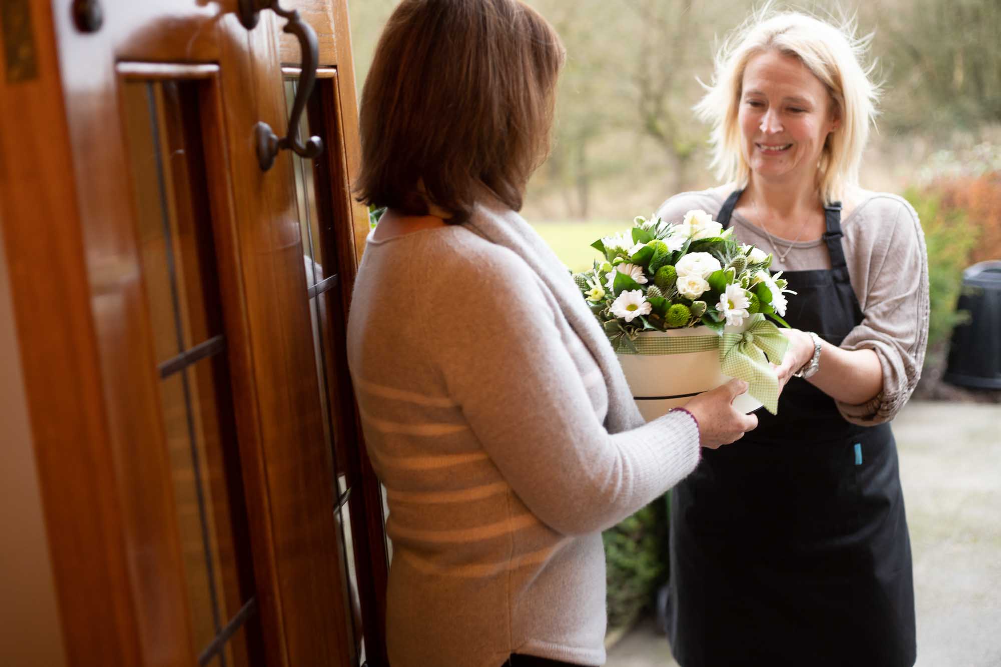 Woman at door receiving a white flower themed hat-box from a florist wearing an apron