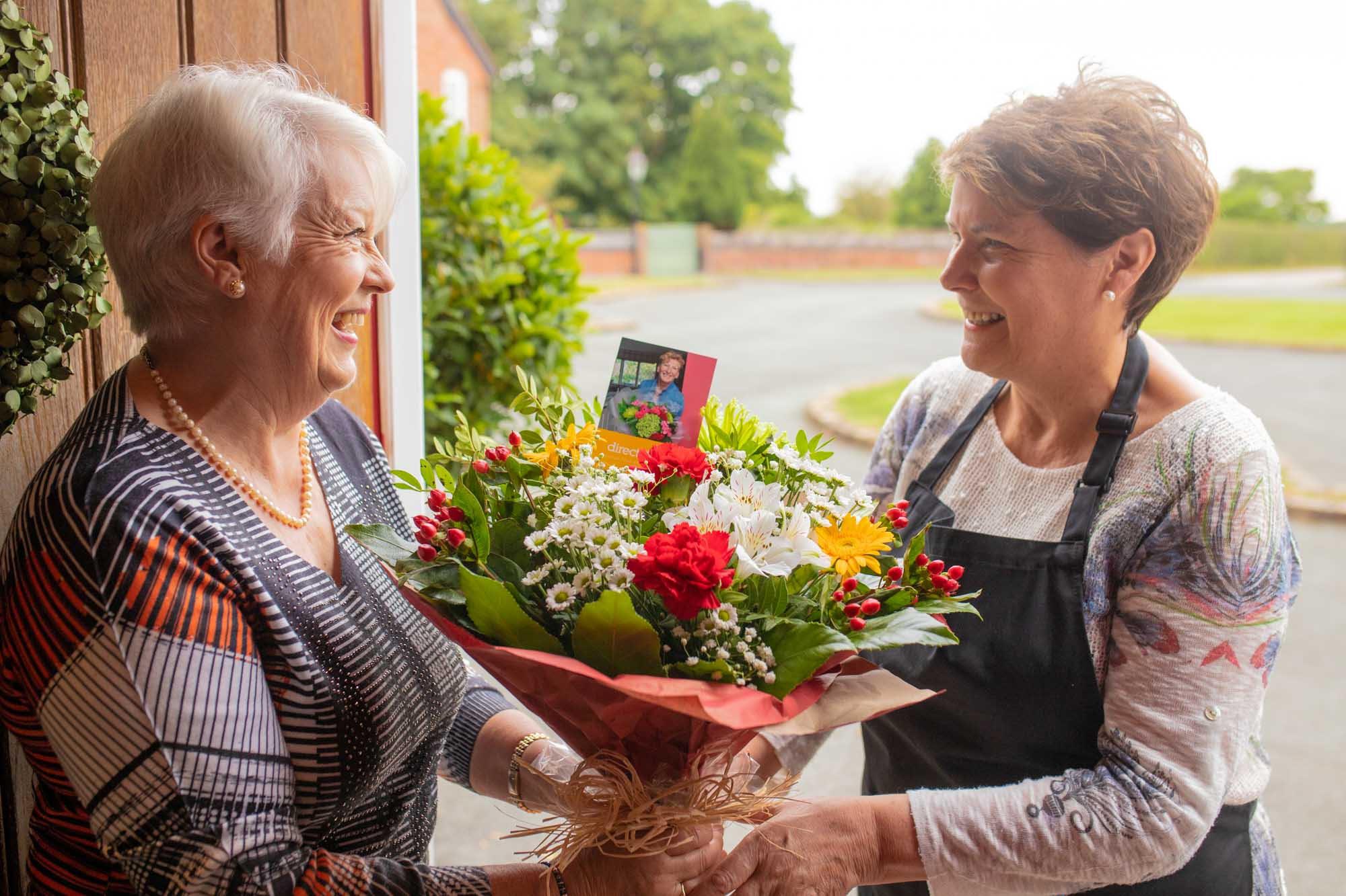 Smiling elderly woman standing at her front door holding a colourful bouquet of flowers, with a festive wreath on the door behind her