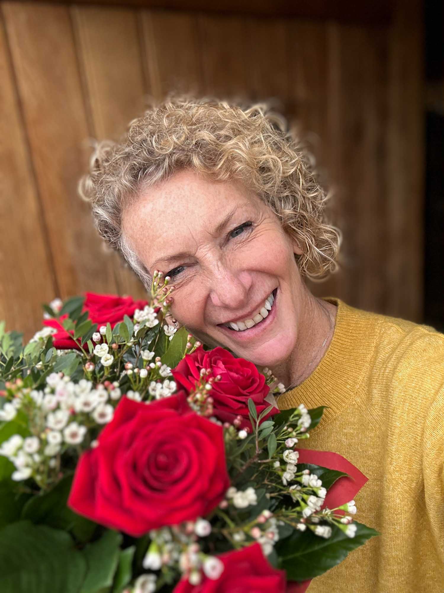 Selfie of a  happy woman wearing a yellow jumper at her front door hugging vibrant red roses.