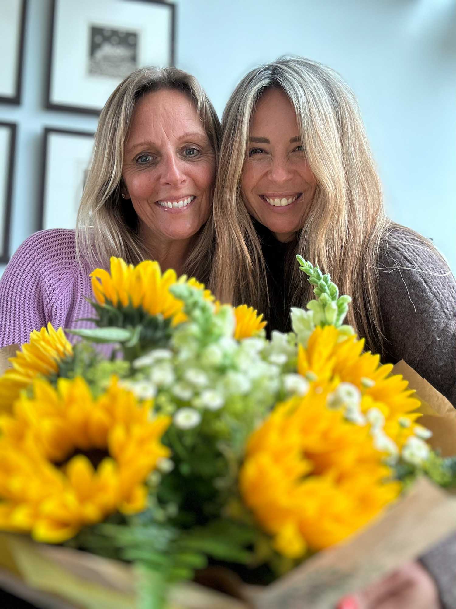 Two happy women with a bokeh sunflower arrangement in the foreground