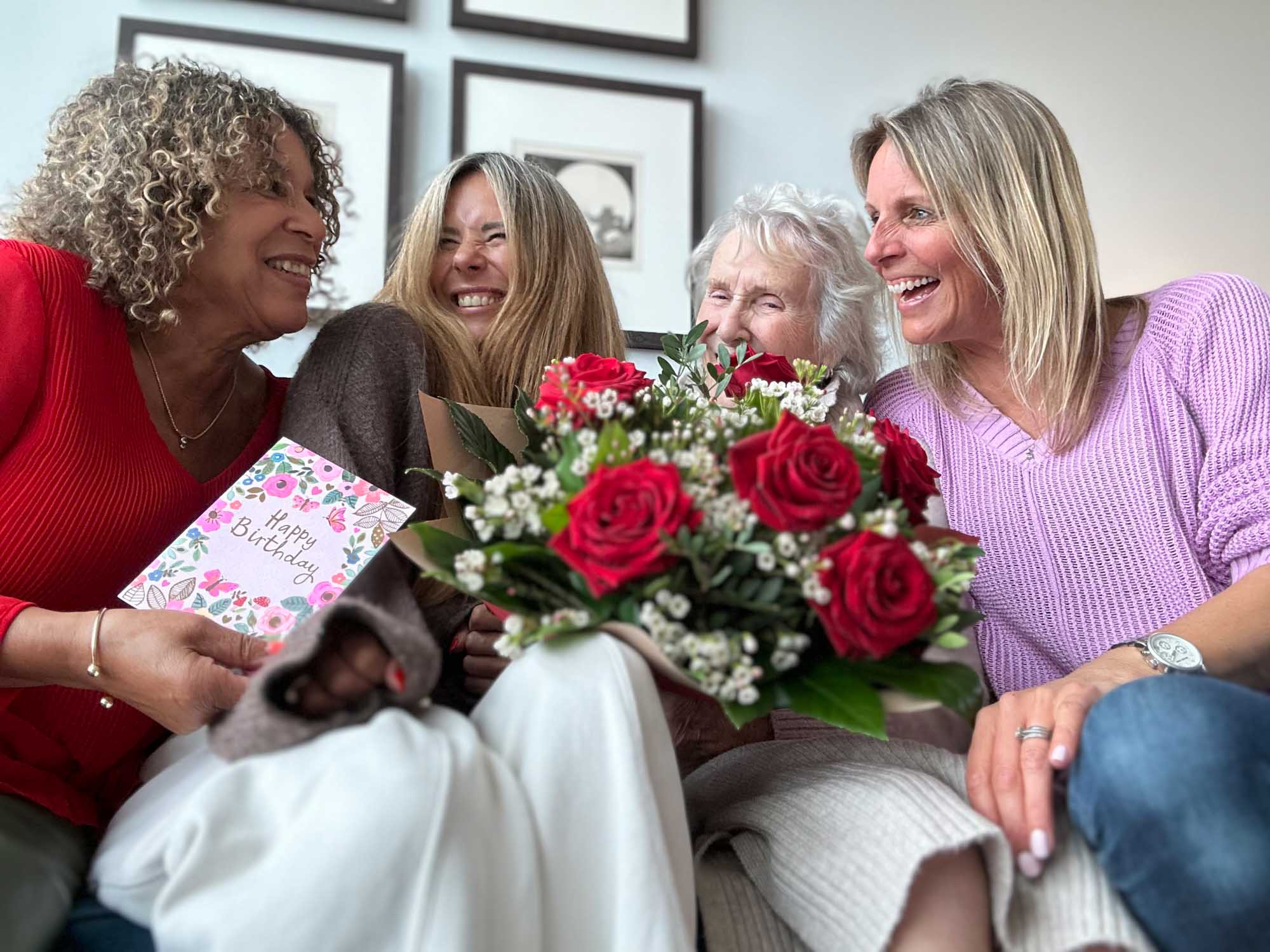 Family sitting together on settee with birthday card and bouquet of flowers. Everyone smiling.