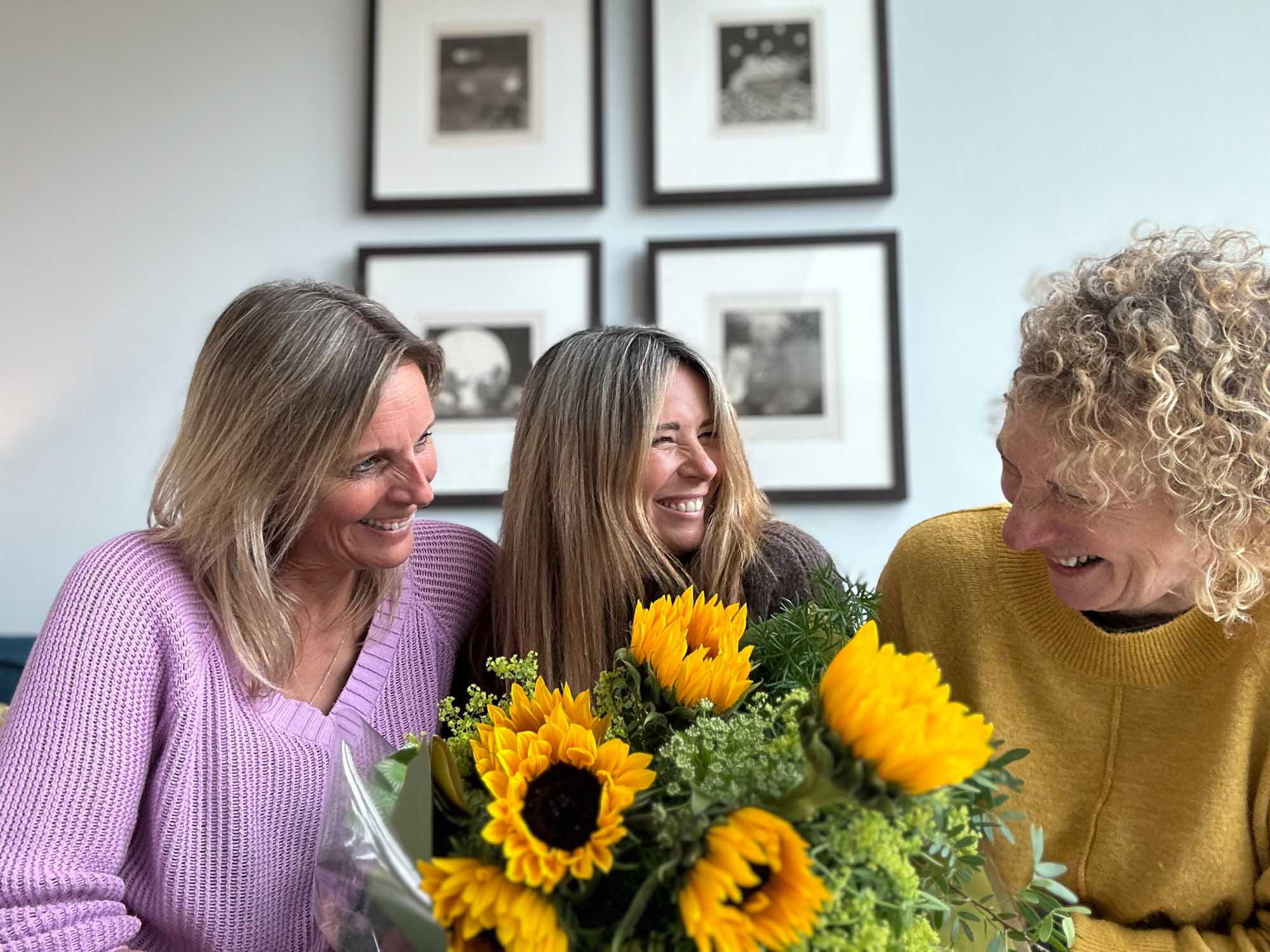 Three friends smiling on sofa while looking at bouquet of sunflowers